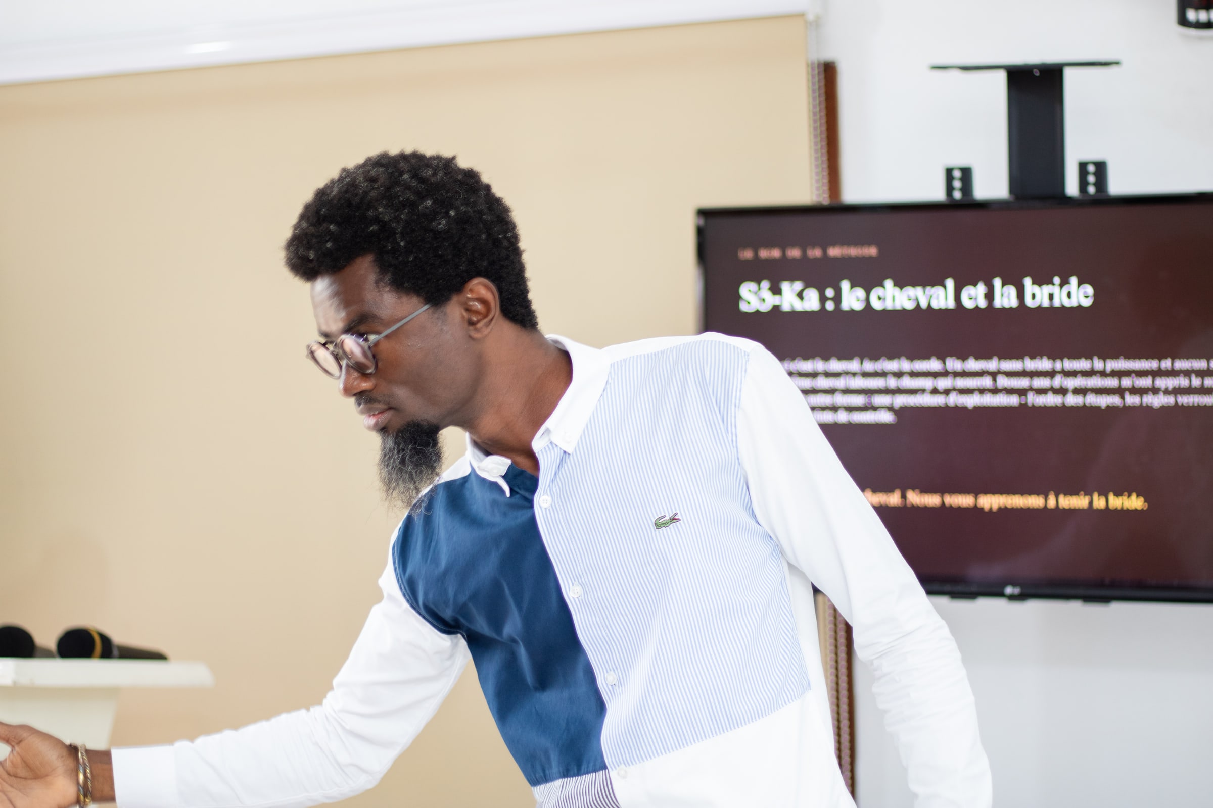 Le fondateur, penché et le bras tendu, devant l’écran qui affiche en entier la diapositive « Sɔ-Ka : le cheval et la bride », pendant la première édition.