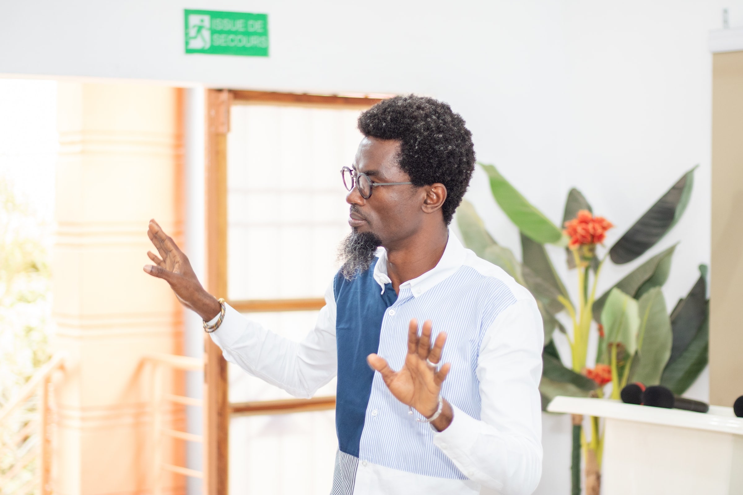 Mohamed Nassirou SOUMANOU, fondateur de Dicko Industries, les deux mains ouvertes en pleine explication pendant la première édition de la Masterclass Dicko Soka ; derrière lui, une porte vitrée baignée de lumière et une plante aux fleurs rouges.